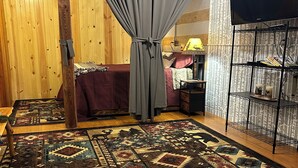 A cozy room with a wooden floor, a patterned rug, a bed with a red bedspread, a grey curtain, and a TV on a black stand. - Cozy, scenic, studio apartment for nature-lovers ! (Paris)