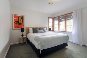 A bedroom with a large bed, a small bedside table, a lamp, a window with blinds, and a framed artwork on the wall.