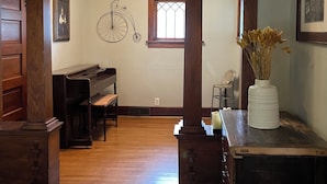 Ein geräumiges Wohnzimmer mit Holzdielen, einem Klavier, einer großen Holzsäule und einer Tischvase mit getrockneten Blumen.
