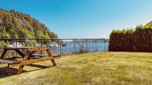 A wooden deck with a metal railing overlooking a body of water, surrounded by grass and a hedge.