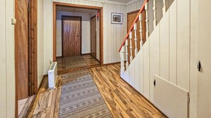 A hallway with wooden flooring, a patterned rug, a staircase with red railings, and a door at the end.