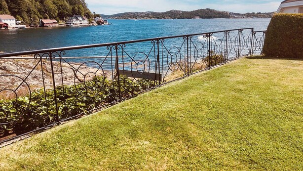 A grassy area with a metal railing overlooking a body of water, with houses and hills in the background.