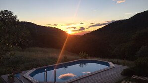 A rectangular swimming pool with a wooden deck, surrounded by grass and trees, with a sunset in the background.