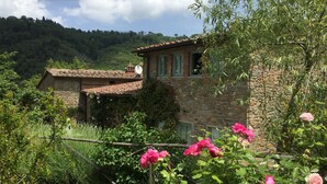 A stone house with a tiled roof, surrounded by lush greenery and blooming pink flowers.