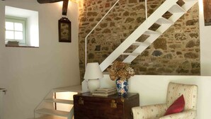 A staircase with a stone wall, a white chair, a small table with a vase and books, and a window with a green door.