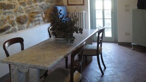 A stone-walled dining area with a long table, chairs, a chandelier, and a door with a view.