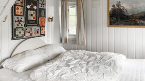 A bedroom with a white bed, a quilt with various patterns, a painting of a landscape, and a window with curtains.