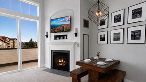 A modern living room with a fireplace, a dining table with benches, a TV mounted above the fireplace, and a cityscape view through large windows. - SL 407: Luxury Remodel | Village Studio with Spectacular Sunset Views (Snowshoe)