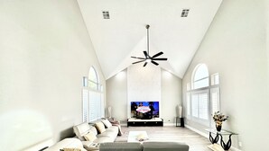 Un salon spacieux avec un plafond haut, un grand téléviseur à écran plat, un canapé d’angle gris et une table basse en verre.