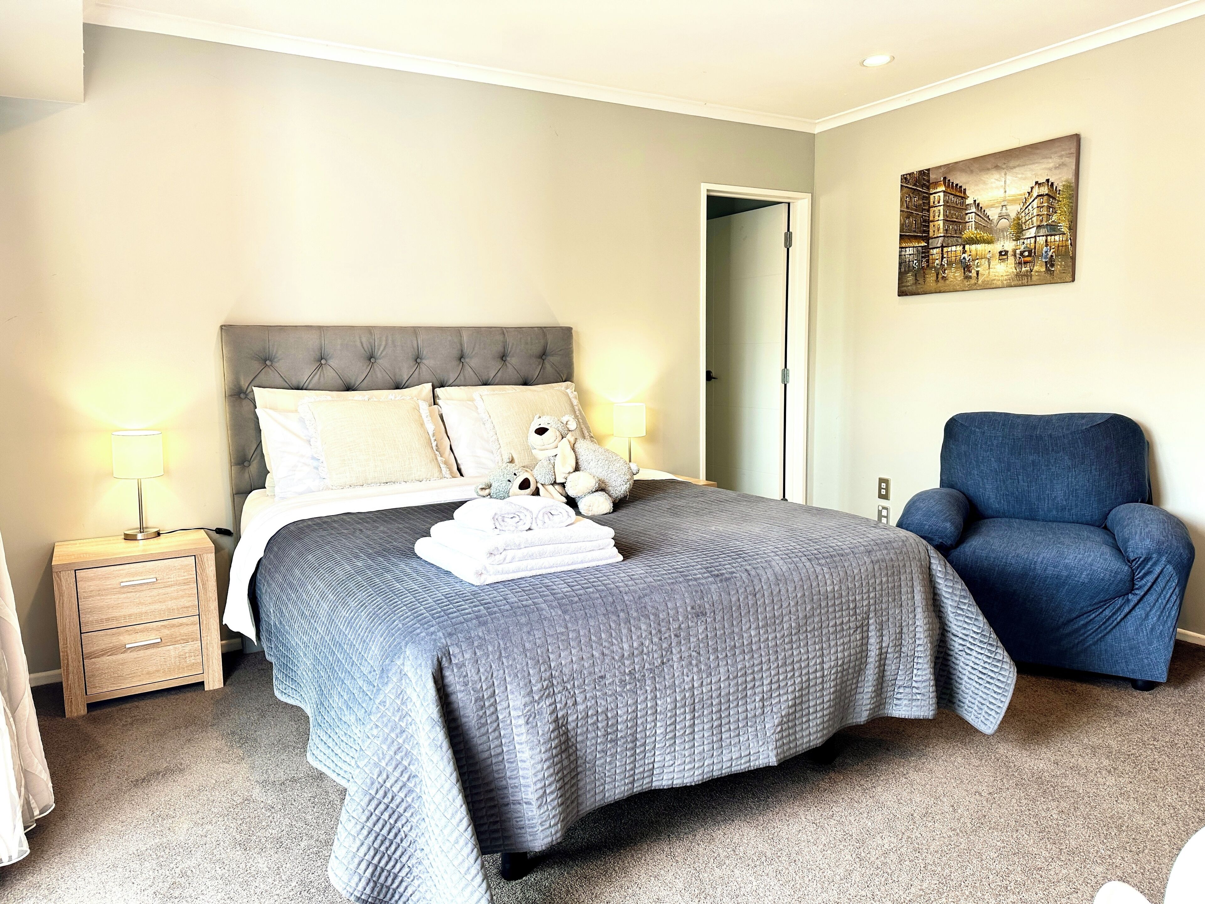 Cama bem arrumada com colcha cinza, uma poltrona azul e um quadro com uma vista urbana na parede.