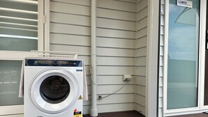 Een wasmachine met voorlaadklep op een veranda, met een witte buitenmuur, een glazen deur en een zichtbaar stopcontact.