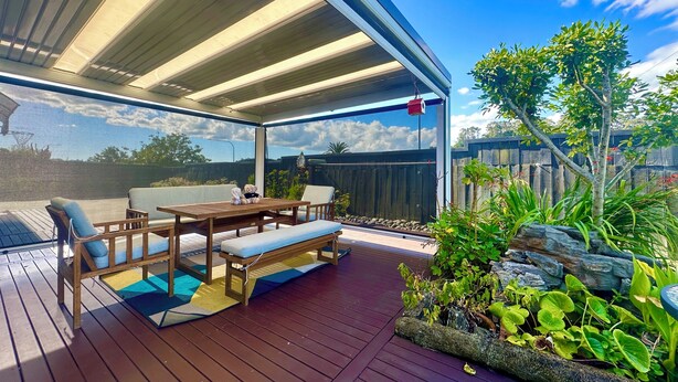Een terras buiten met een houten dek, een overdekte zithoek en uitzicht op een achtertuin met houten omheining en begroeiing.
