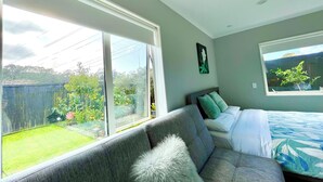 Una camera da letto con una grande finestra, un divano grigio, un letto con copriletto a fantasia blu e bianca e una pianta verde sul davanzale.