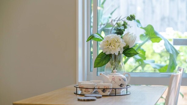 Un tavolo di legno con un bollitore, tazze e piattini, e un vaso con fiori vicino a una finestra.