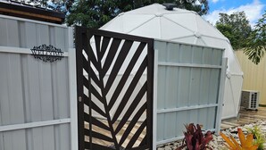 Een omheind gebied met een 'WELCOME' bord, een witte structuur met een koepeldak, en een tuin met diverse planten.