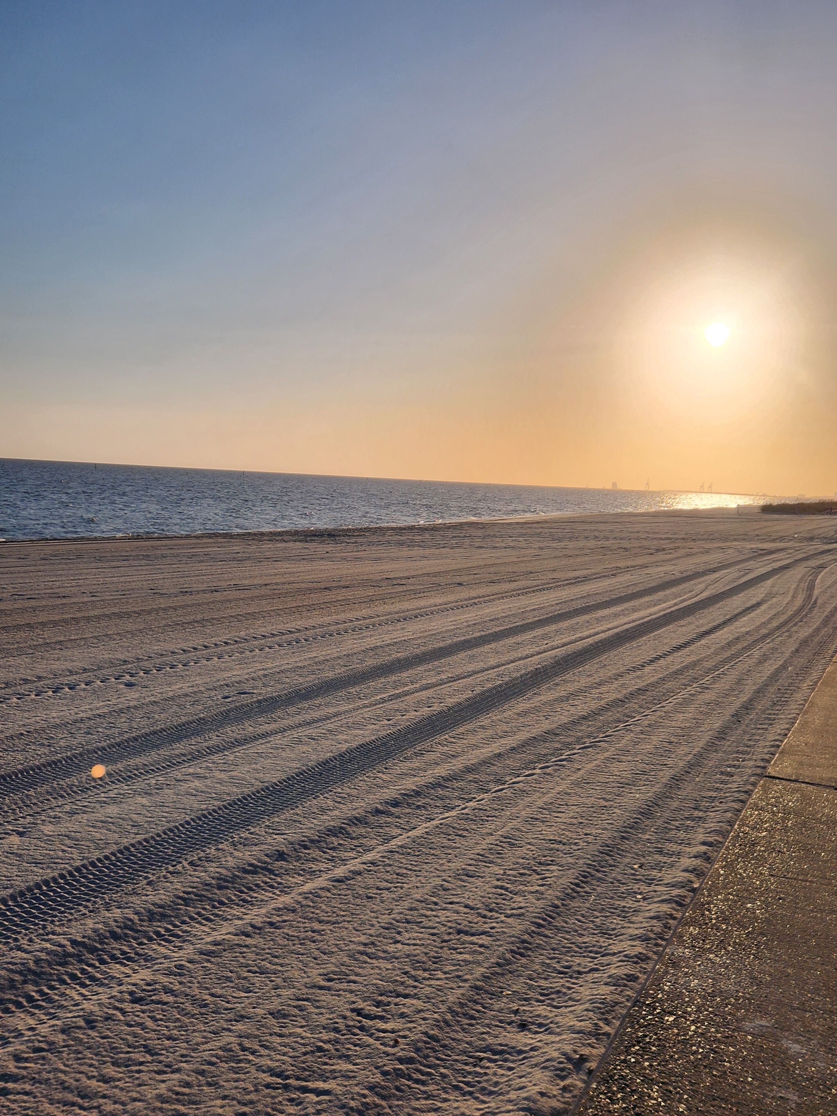 Een strand met sporen van banden die naar het water leiden, een heldere lucht en de zon die ondergaat of opkomt.