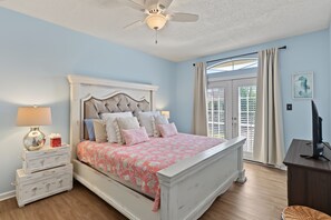 Un dormitorio con cama, mesitas de noche, lámpara, ventilador de techo, ventana con cortinas e imagen enmarcada en la pared.