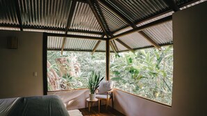 A room with a wooden floor, a bed, a chair, a small table with a plant, and a large window with a view of greenery.