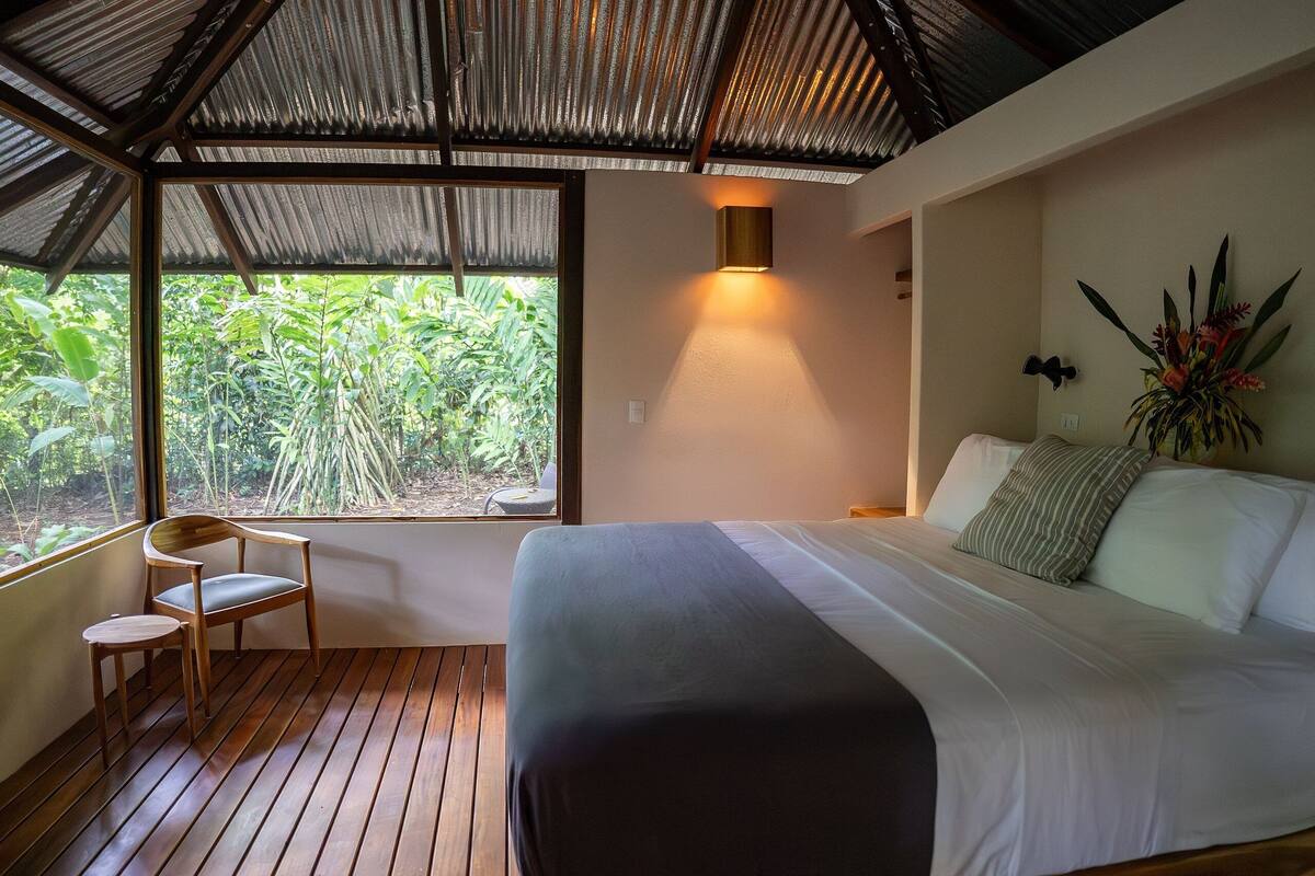 Une chambre à coucher comprenant un lit, une chaise, une fenêtre donnant sur un espace verdoyant et un luminaire fixé au mur.