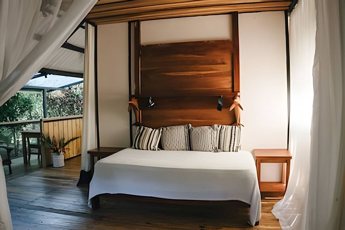 Une chambre à coucher avec un grand lit, une tête de lit en bois, deux tables de chevet et une vue sur l’extérieur.