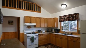 A compact kitchen with wooden cabinets, a stove, a sink, and a window with a view of trees. - The Moose Nugget Cabin (Trapper Creek)