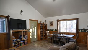 A cozy living room with a television, bookshelf, dining table, and a large window. - The Moose Nugget Cabin (Trapper Creek)