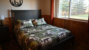 A bedroom with a wooden headboard, a quilted bedspread featuring animal prints, a nightstand with a lamp, and a window with a view of trees. - The Moose Nugget Cabin (Trapper Creek)