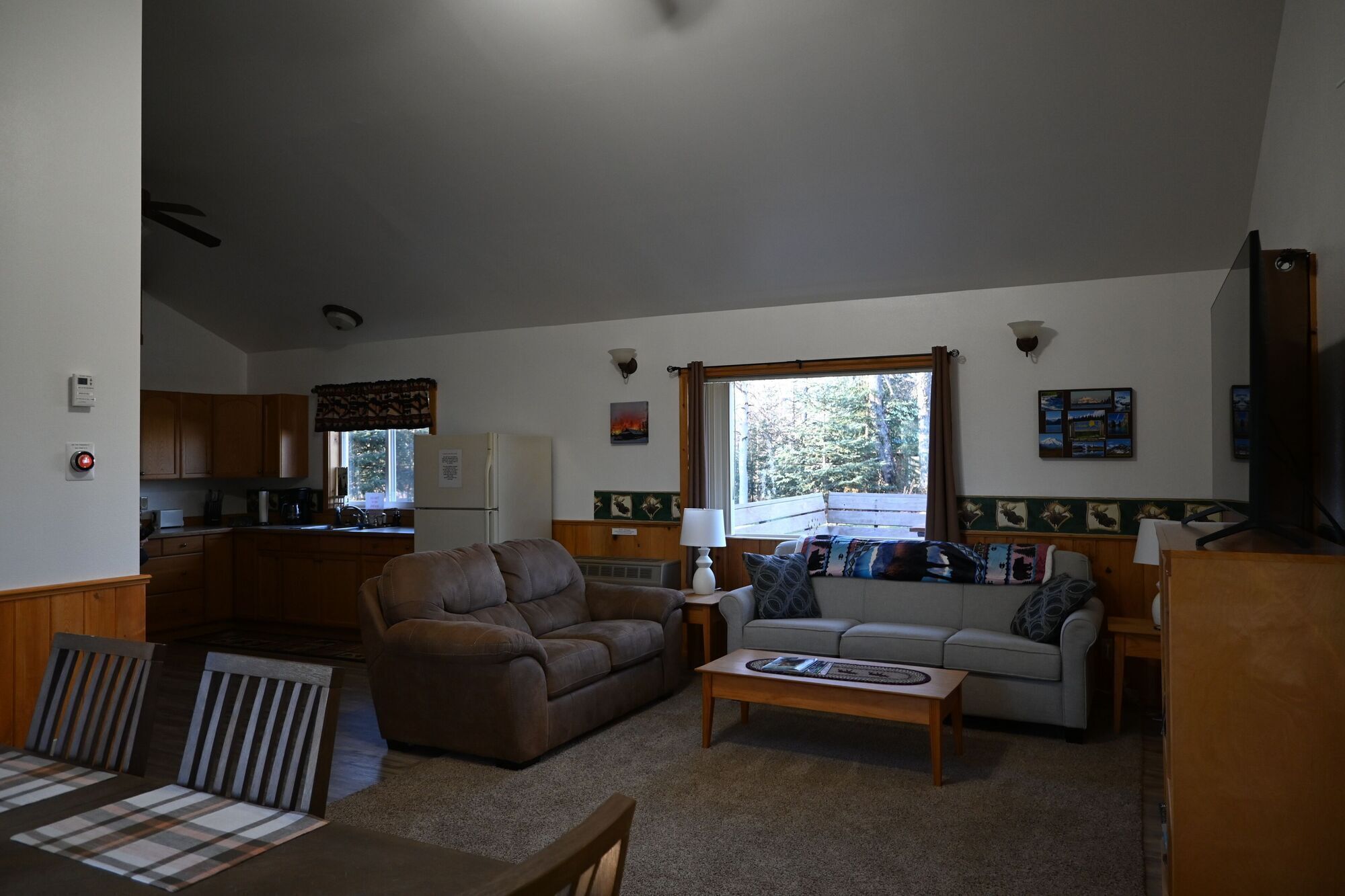 A cozy living room with a sofa, a coffee table, and a dining table with chairs. There is a kitchen area with wooden cabinets and a refrigerator.