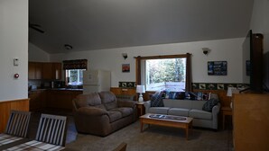 A cozy living room with a sofa, a coffee table, and a dining table with chairs. There is a kitchen area with wooden cabinets and a refrigerator. - The Moose Nugget Cabin (Trapper Creek)