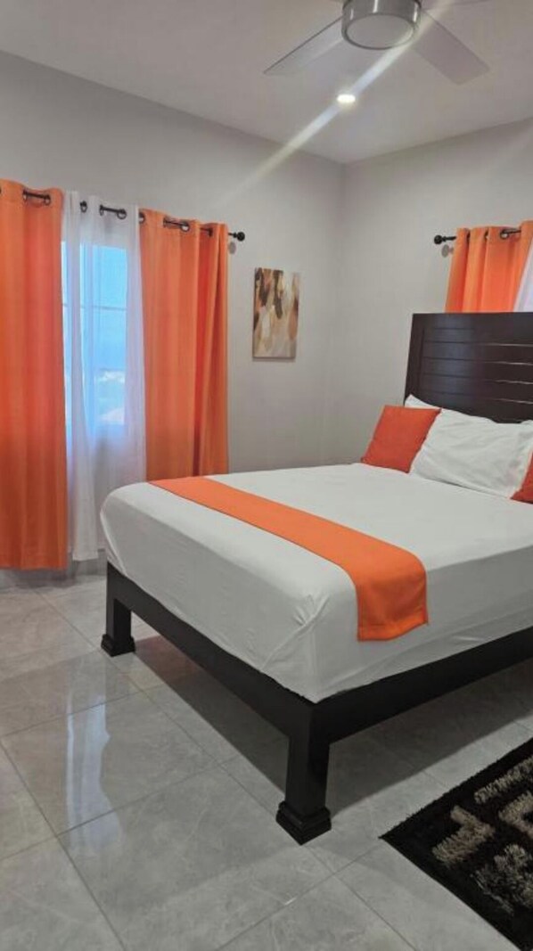 A bedroom with a bed, orange curtains, a ceiling fan, and a painting on the wall.