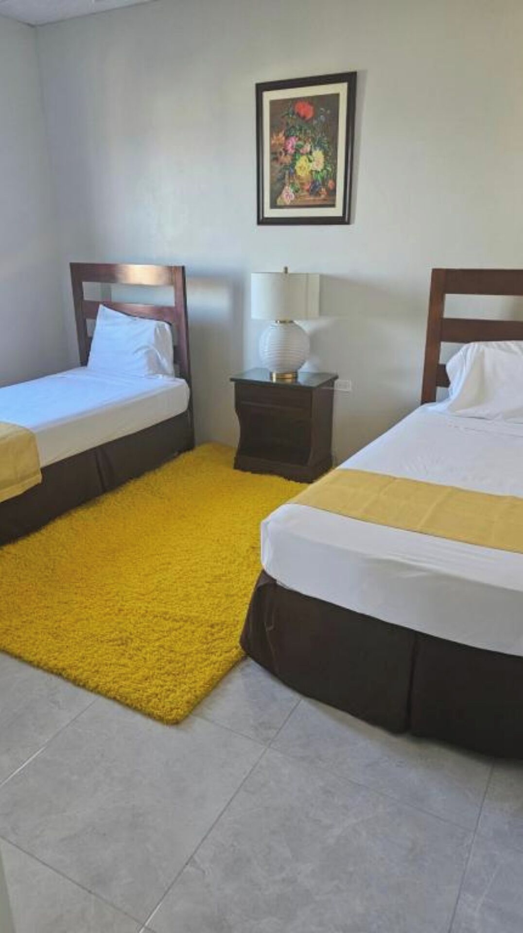 Quarto de hotel com duas camas de solteiro, uma pequena mesa de cabeceira com abajur, um tapete amarelo e um quadro na parede.