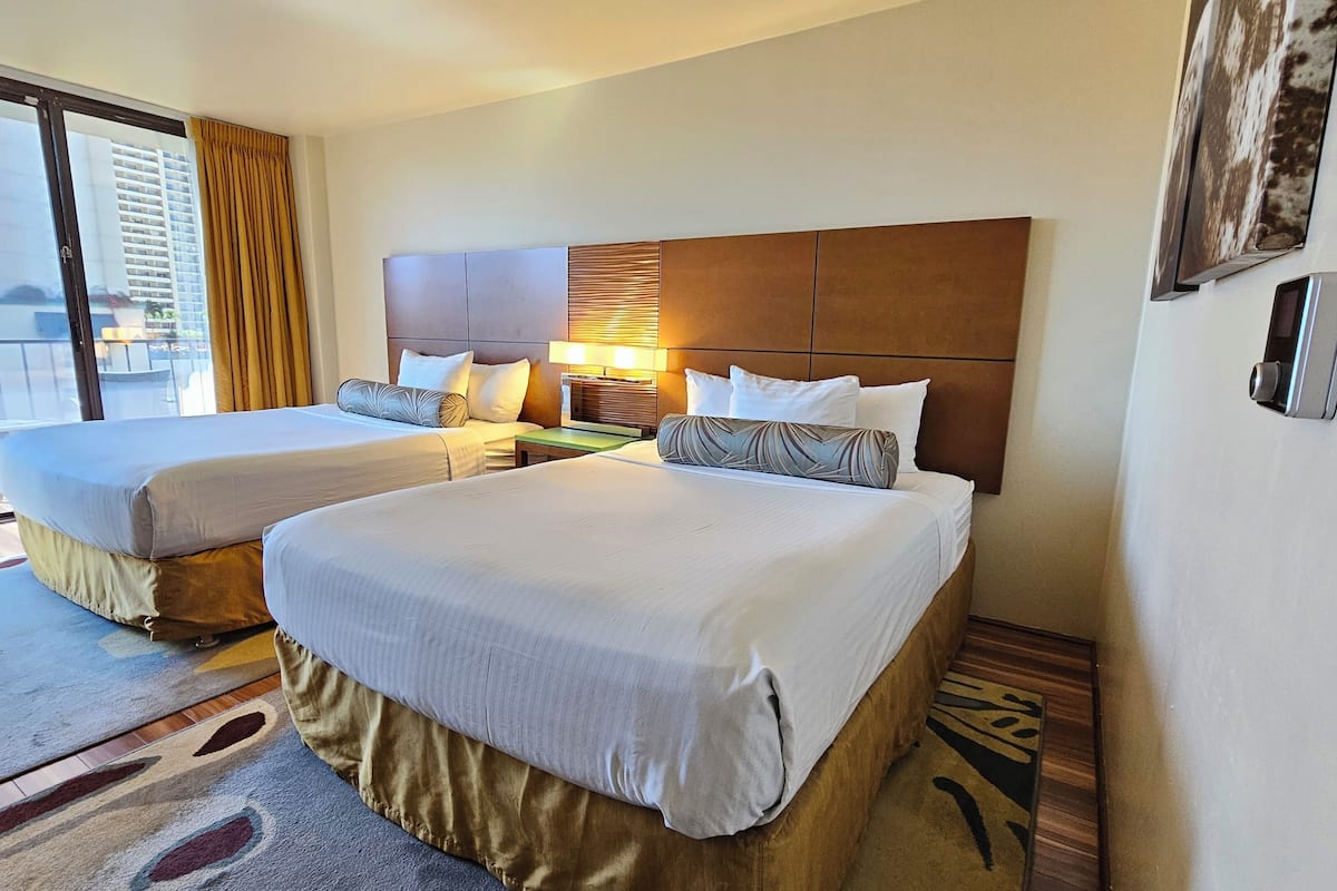 Quarto de hotel com duas camas, uma janela grande com cortinas, cabeceira de madeira e vista para a cidade.