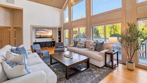 A spacious living room with a large sofa, a coffee table, and a TV mounted on the wall. There are large windows providing natural light and views of the outdoors. - Lakeside Escape – Beach Access, Heated Pool, Hot Tub & Sunset Views (Lakeside)