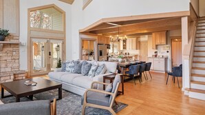 A spacious living area with a sofa, coffee table, and a dining table with chairs. A kitchen is visible in the background with wooden cabinets and a staircase leading to an upper level. - Lakeside Escape – Beach Access, Heated Pool, Hot Tub & Sunset Views (Lakeside)