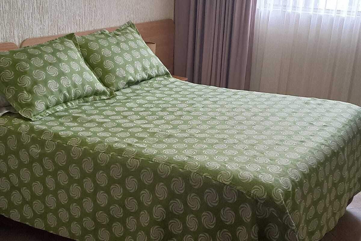 A bed with a green patterned bedspread and pillows, a wooden headboard, and a window with curtains.