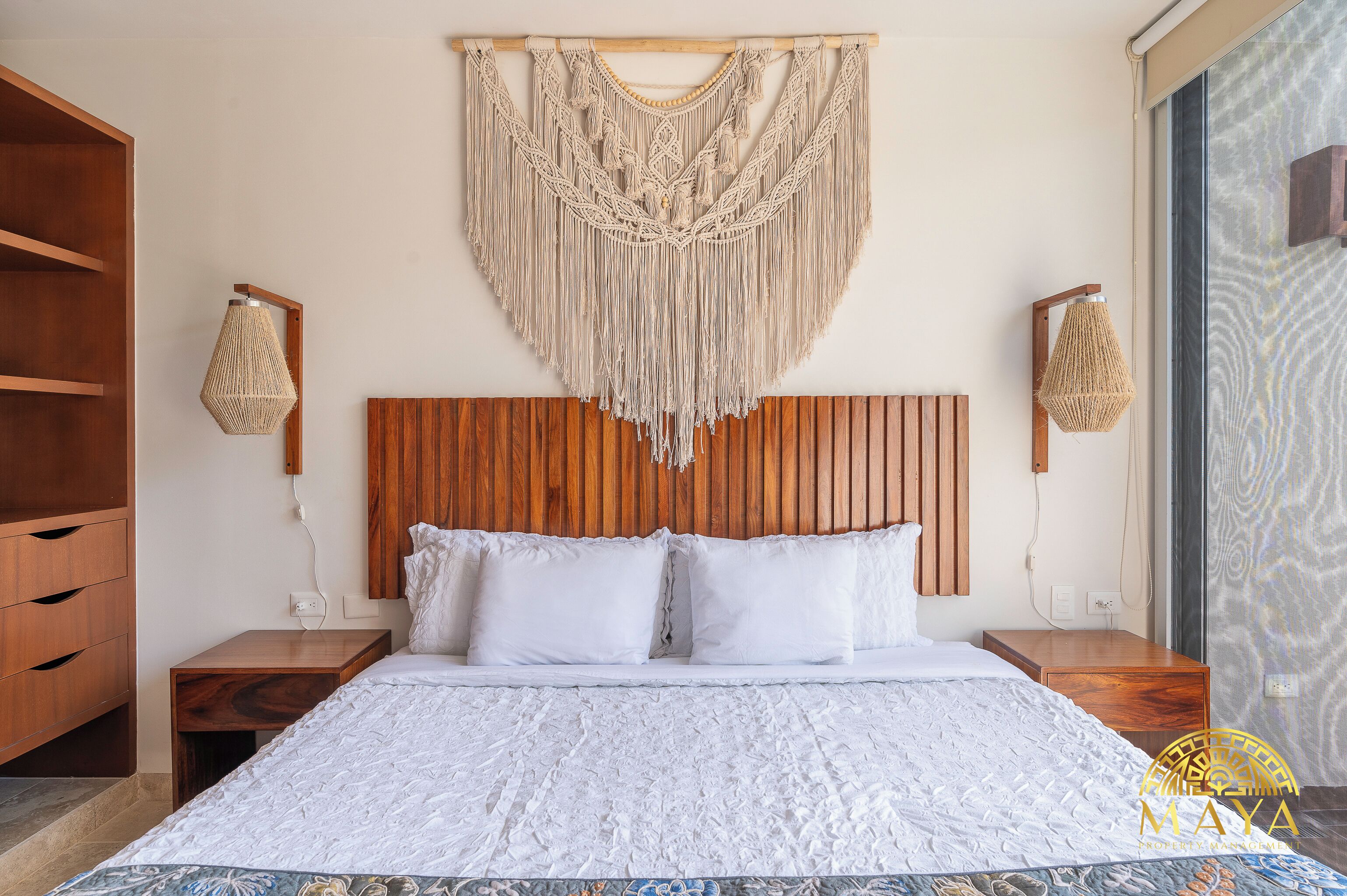 Une chambre à coucher avec une tête de lit en bois, une décoration murale en macramé, deux tables de chevet avec des lampes et une couvre-lit à motifs.