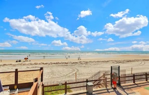 Uitzicht op het strand vanaf een houten terras met een leuning, een heldere lucht en een zandstrand met mensen en parasols.