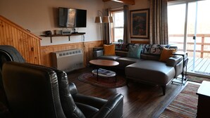 A cozy living room with a leather chair, a sofa with patterned cushions, a flat-screen TV, and a large window with curtains. - The Denali View Cabin (Trapper Creek)