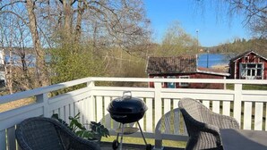 A covered deck with wicker furniture, a barbecue grill, and a view of a red building and trees. - 4 Star Holiday Home in Gnesta (Gnesta)