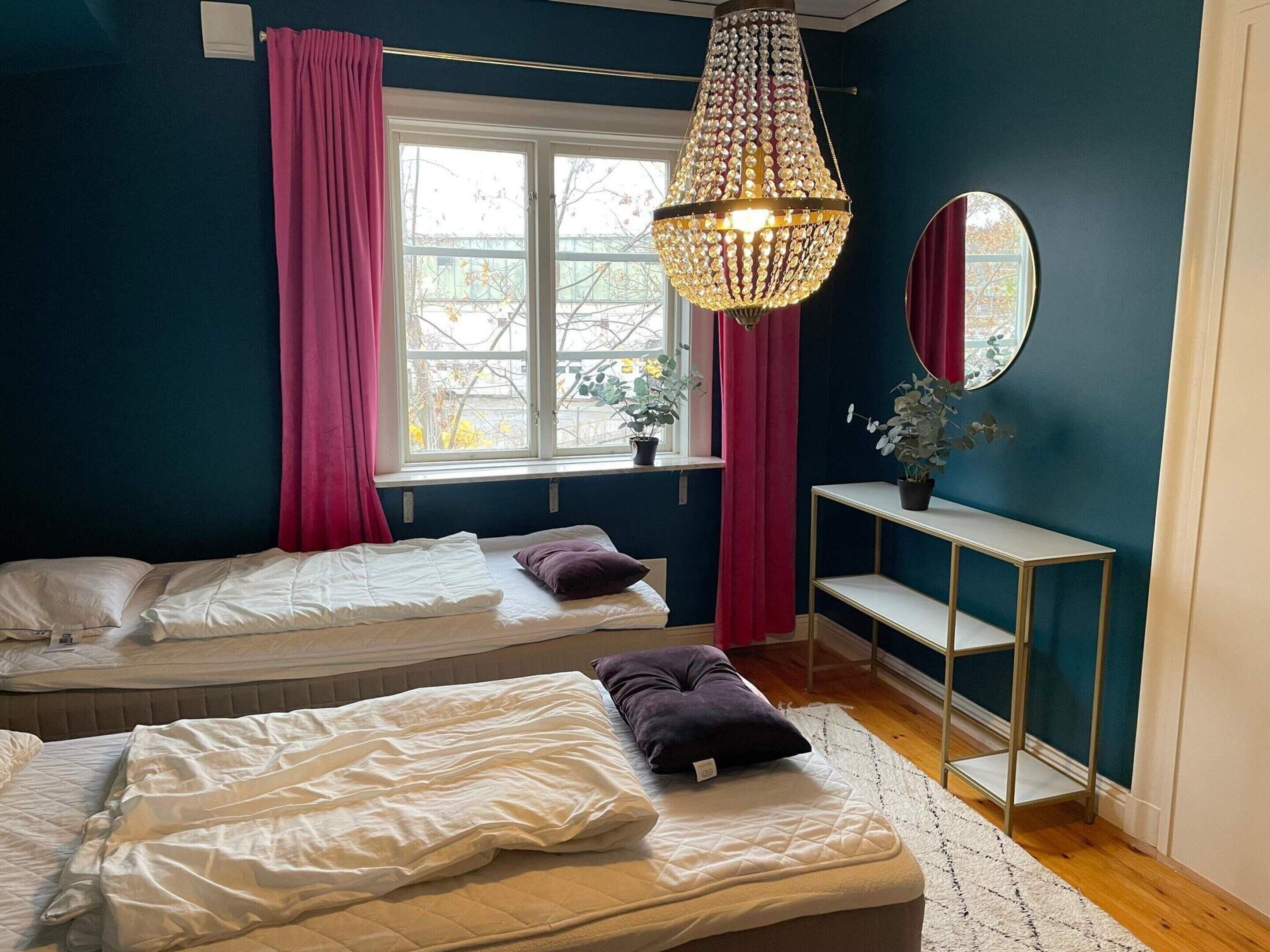 A bedroom with two beds, a large window with curtains, a chandelier, a mirror, and a small table with a plant.