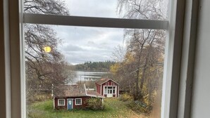 A view through a window of a small house with a red roof and white trim, surrounded by trees and a body of water. - 4 Star Holiday Home in Gnesta (Gnesta)