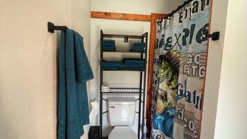 Ein kleines Badezimmer mit einer weißen Toilette, einem Handtuchhalter mit einem grünen Handtuch, einer schwarzen Ablage mit gefalteten Handtüchern und einem Duschvorhang mit Text.