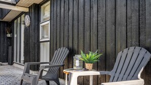 Two plastic chairs and a small table with a potted plant on a cobblestone pavement in front of a wooden building with vertical siding.