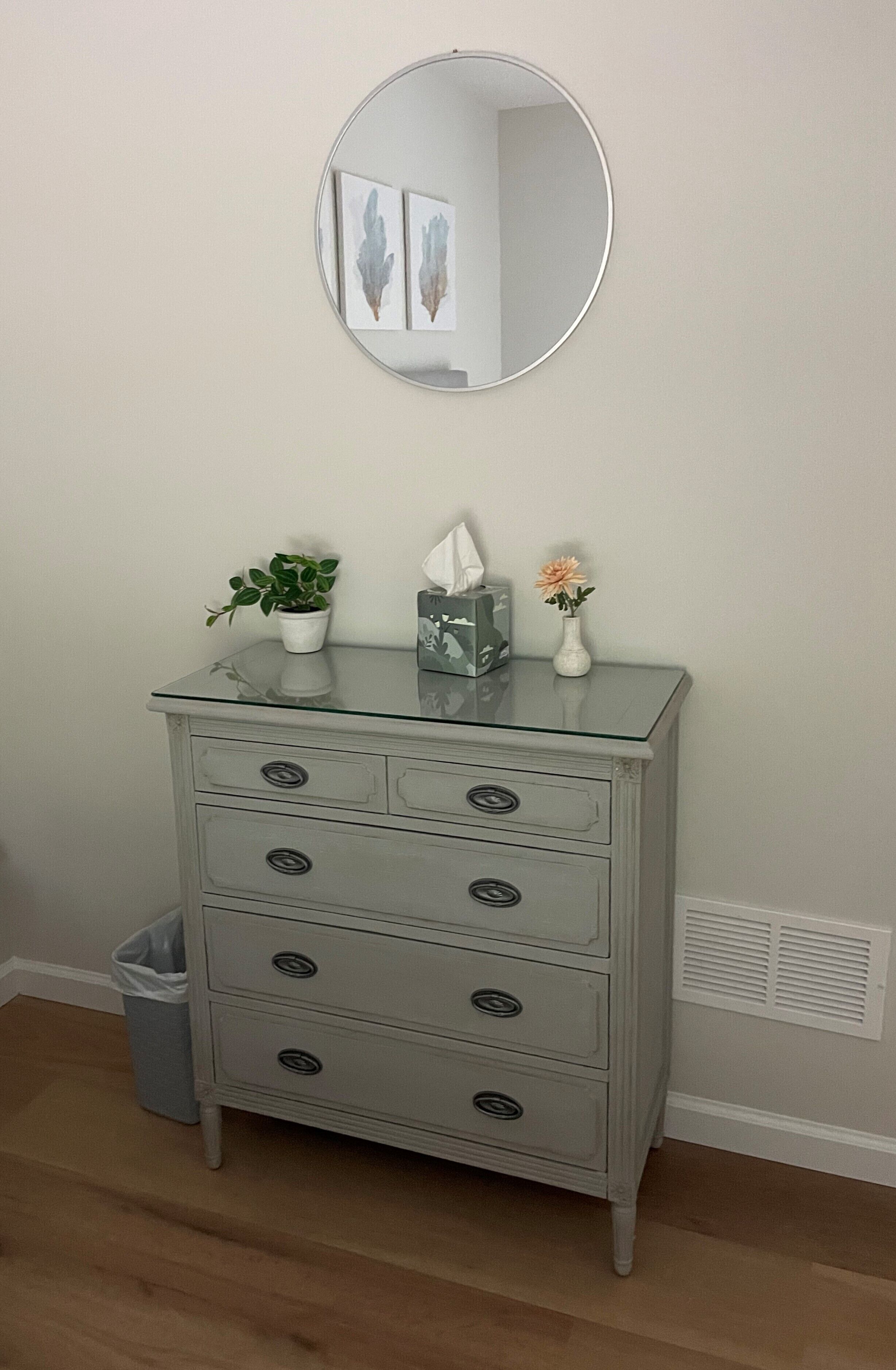 A white dresser with a glass top, a round mirror above it, and a potted plant on top.