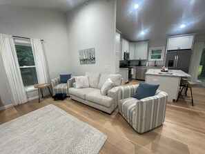 A modern living room with a sofa, armchair, and a small table. A kitchen is visible in the background with white cabinets and a refrigerator.