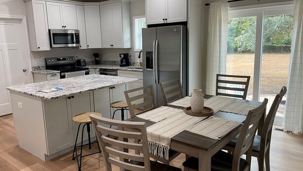 A modern kitchen with white cabinets, a granite countertop, and stainless steel appliances. There is a dining area with wooden chairs and a table, and a view of a garden through a sliding glass door.