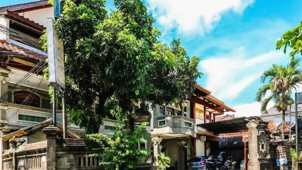 A street with traditional buildings, a parked car, and a tree in the foreground. - Hotel Candra Adigraha (Denpasar)