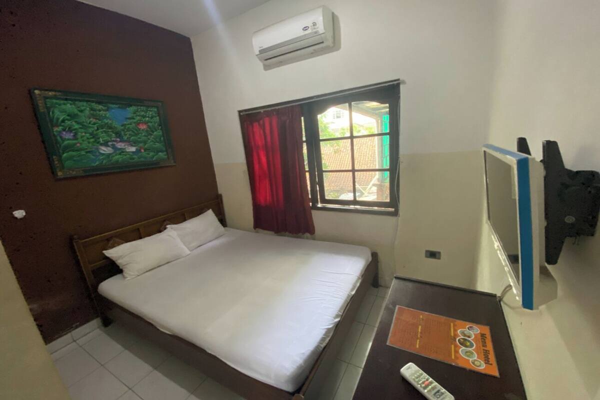A hotel room with a single bed, an air conditioner, a window with curtains, a desk with a remote control, and a wall-mounted TV.