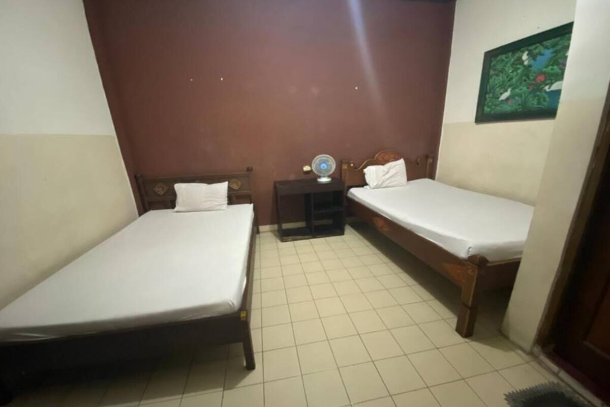 A small, clean room with two single beds, a nightstand with a fan, and a small table.
