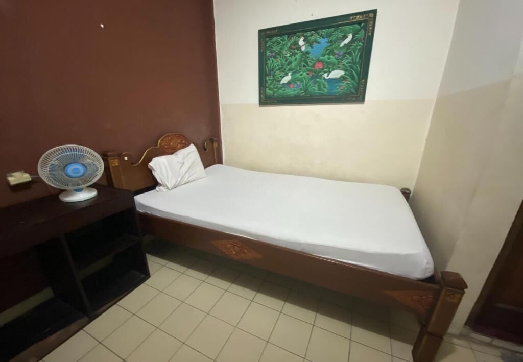 A small, single-bed room with a wooden headboard, a white mattress, a wall-mounted fan, and a framed picture above the bed.